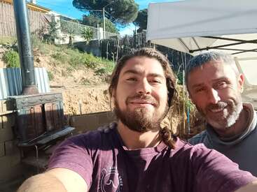 Das Bild zeigt zwei Männer, die für ein Selfie vor einem weißen Vordach posieren, mit einem Holzofen und einem Berghang im Hintergrund.