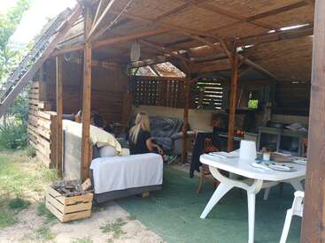 Un refugio rústico de madera al aire libre con sofás, gente descansando, una mesa de plástico blanco, sillas, una alfombra verde y un ambiente acogedor, rodeado de vegetación y luz natural.
