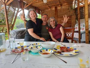 Tres personas sonrientes están sentadas juntas a la mesa después de comer, disfrutando de la compañía mutua en un acogedor espacio exterior de madera bañado por la luz natural.