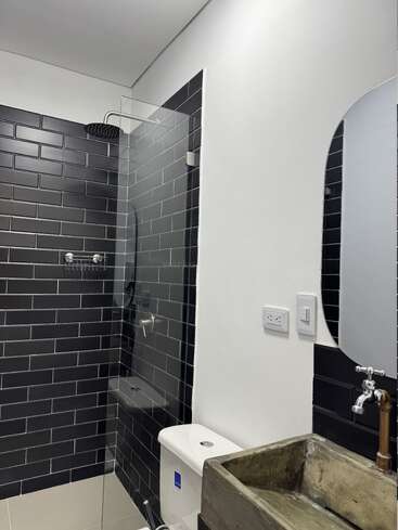 Este cuarto de baño cuenta con azulejos negros, una ducha acristalada, un moderno cabezal de ducha tipo lluvia, un lavabo rústico de hormigón, un inodoro, un espejo minimalista y paredes blancas para crear contraste.