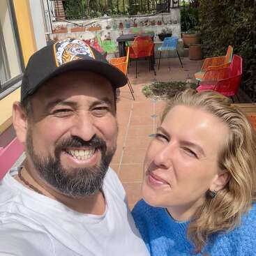 Un hombre y una mujer sonrientes posan para un selfie al aire libre en un día soleado. Detrás de ellos, hay sillas de colores, mesas, plantas y un patio embaldosado.