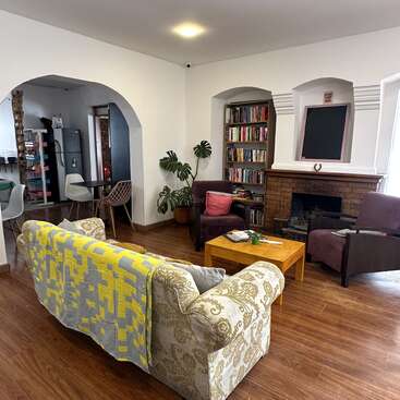 Este acogedor salón cuenta con un sofá estampado, una manta amarilla, sillones, mesa de centro, estantería, chimenea, suelo de madera, plantas de interior y arcos de entrada a la cocina.