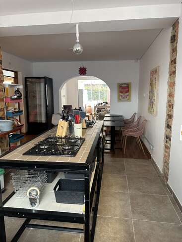 A modern kitchen with a large island stovetop, utensils, and storage baskets. Dining area with pink chairs visible through arched doorway, brick and bright decor accents.