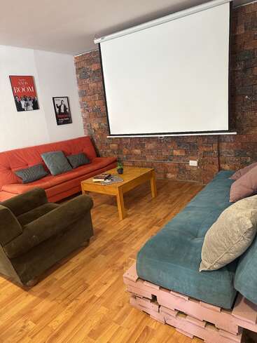 Cozy living room with wooden floor, red, green, and teal couches, coffee table, projector screen, brick accent wall, and two framed movie posters. Inviting atmosphere.