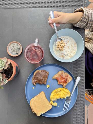 A breakfast scene with cereal in milk, tea, and a blue plate holding toast with peanut butter, chocolate spread, jam, and scrambled eggs. Succulent plant nearby.