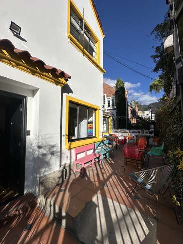 A sunny patio with colorful chairs and benches sits beside a white building with yellow trim. Potted plants, blue sky, and shadows create a welcoming atmosphere.