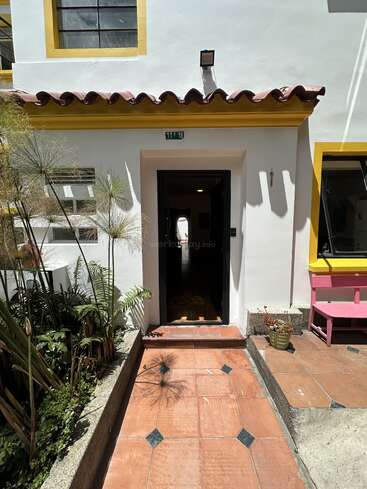 A white house entrance with a tiled walkway, yellow trim around windows, potted plants, a pink bench, and sunlight casting shadows onto the pathway and wall.