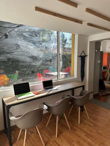 A bright workspace features three modern chairs, a long desk, two laptops, notebooks, large windows with outdoor view, and cozy natural light on wooden floors.