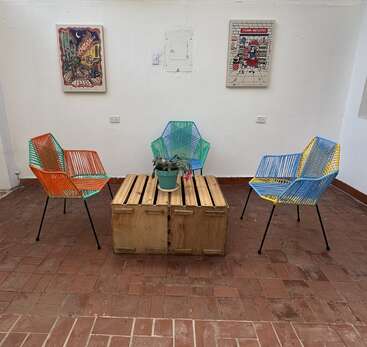 This image shows a cozy seating area with three colorful chairs around a wooden table, a potted plant centerpiece, art on white walls, and tiled floor.