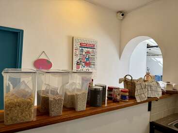 Clear containers of cereal and oats, jars of peanut butter and jam, a coffee canister, and baskets on a wooden shelf in a bright, cozy kitchen.