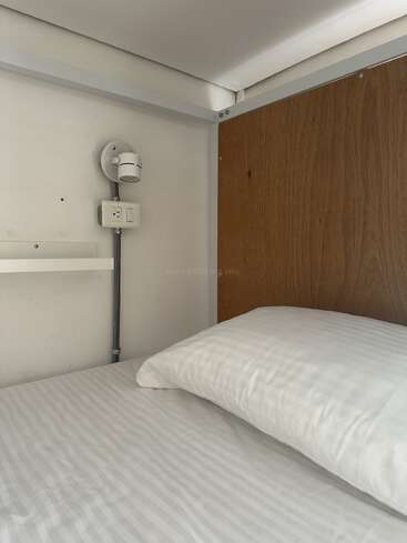 This image shows a minimalistic bunk bed setup with a wooden panel, a single white pillow, striped bedding, a spotlight lamp, and a small white shelf.