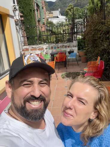A smiling man and woman pose for a selfie outdoors on a sunny day. Behind them, there are colorful chairs, tables, plants, and a tiled patio.