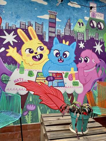 A colorful mural features cartoon animals playing a board game, set against a vibrant cityscape and mountains. Bright chairs and a potted plant complete the scene.