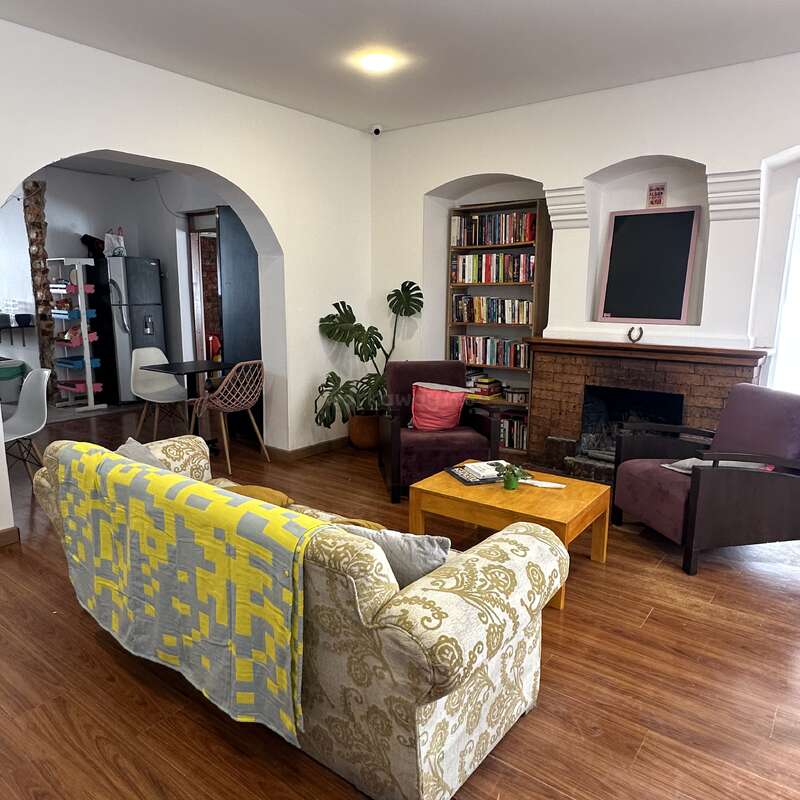 Este acogedor salón cuenta con un sofá estampado, una manta amarilla, sillones, mesa de centro, estantería, chimenea, suelo de madera, plantas de interior y arcos de entrada a la cocina.