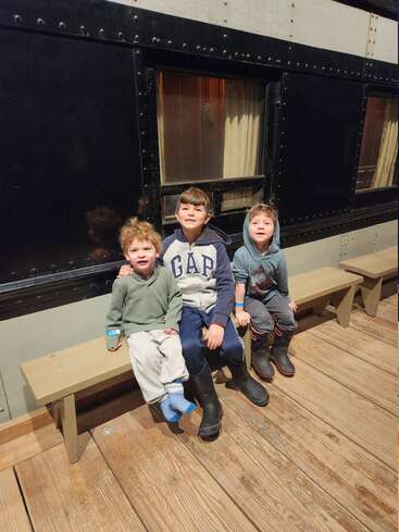 Three young boys sit together on a wooden bench, smiling, in front of an old-fashioned train car with black paneling and curtained windows, indoors, warmly dressed.
