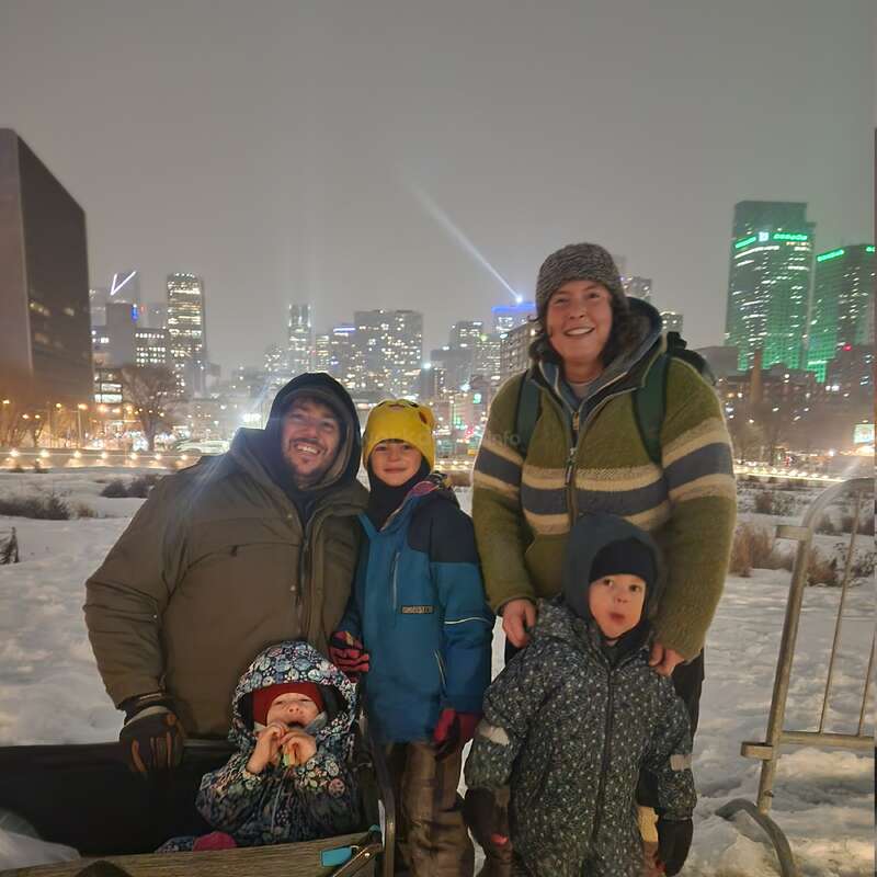 A family of five, bundled in winter clothing, poses happily together in a snowy park at night, with a brightly lit city skyline behind them.