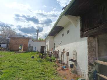 This image shows a rustic courtyard with a whitewashed building, garden tools, potted plants, a grassy yard, wooden gate, and cloudy sky overhead, creating a peaceful rural scene.