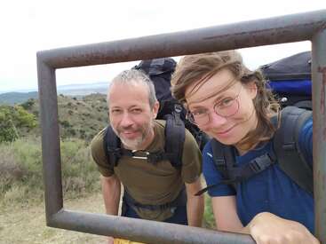 A man and a woman with backpacks smile through a rusty metal frame, standing on a scenic hillside with mountains and greenery in the background, enjoying hiking.