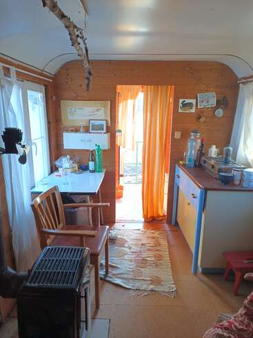This cozy wooden interior features a small kitchen, stove, table, chair, and orange curtain. Natural light fills the room, creating a warm, inviting atmosphere.