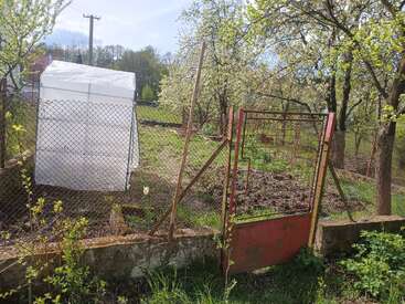 Un petit jardin entouré d'une clôture grillagée et d'un portail rouge, avec une serre blanche, des arbres en fleurs, de l'herbe verte et un temps ensoleillé, clair et paisible.