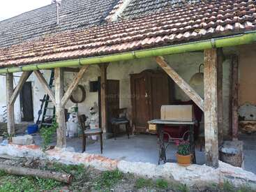 Cette image montre un porche rustique avec des poutres en bois, de vieux meubles, une armoire, des chaises, une machine à coudre, des outils éparpillés, une plante en pot et un toit en tuiles usé par les intempéries.