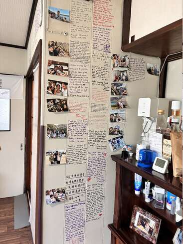 Cette image montre un mur orné de messages écrits à la main et de nombreuses photos de groupes de personnes souriantes, immortalisant des souvenirs et des mots sincères dans une pièce chaleureuse.