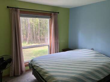 Um quarto aconchegante com paredes verdes e azuis, uma janela grande com cortinas bege, uma colcha listrada, uma mesinha de cabeceira e um ventilador. Vista para a floresta lá fora.