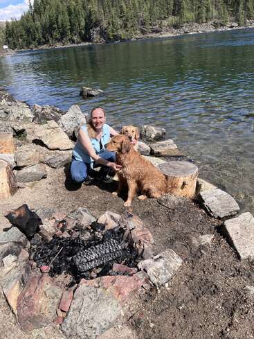 Uma mulher está ajoelhada à beira de um lago com dois golden retrievers, cercada por pedras e uma fogueira. As árvores e a água cristalina criam um cenário tranquilo ao ar livre.