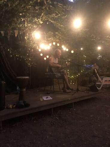 Une personne joue de la guitare sur une petite scène en plein air, la nuit, entourée de guirlandes lumineuses et d'arbres. Ambiance chaleureuse et intime avec un public détendu et des instruments à percussion à proximité.