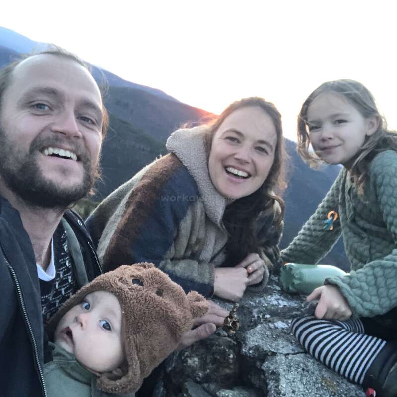L'image représente une famille de quatre personnes posant pour un selfie sur un affleurement rocheux, avec des montagnes en arrière-plan et un ciel lumineux.