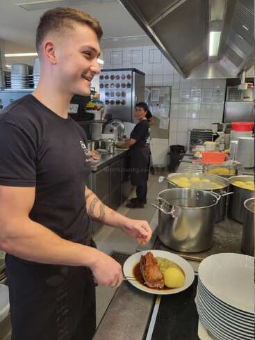 Un jeune chef souriant se tient dans une cuisine animée, tenant une assiette contenant du rôti de porc, de la sauce, de la purée de pommes de terre et des légumes. Des casseroles et du personnel sont visibles derrière lui.