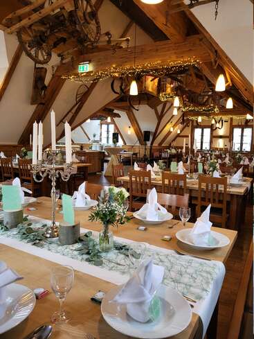Une salle de banquet rustique ornée de poutres en bois, de guirlandes lumineuses et d'outils agricoles anciens. Des tables élégantes sont dressées avec des serviettes blanches, des fleurs et des verres.