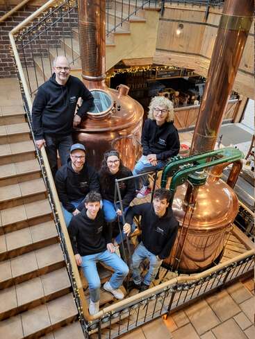 Un groupe de six personnes pose ensemble près de grandes cuves de brassage en cuivre dans une distillerie. Elles portent des sweats à capuche noirs assortis et sourient chaleureusement dans un environnement rustique.
