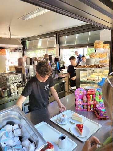 Deux jeunes travailleurs préparent le café et les desserts dans un café très fréquenté. Des tranches de gâteau, du café et des friandises colorées sont visibles, tandis que les clients attendent au comptoir.