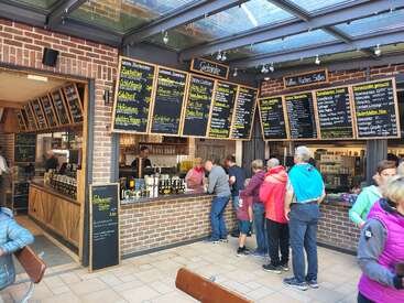 Cette image montre un comptoir de restauration allemand très animé, avec de grands menus affichés à la craie. Les gens font la queue pour commander. Le cadre est lumineux, décontracté et accueillant.