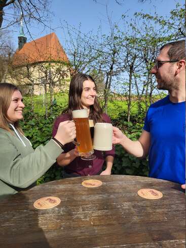 Trois amis trinquent joyeusement avec de grandes chopes de bière à une table en bois en plein air, profitant d'une journée ensoleillée. La verdure et un bâtiment pittoresque se trouvent à l'arrière-plan.