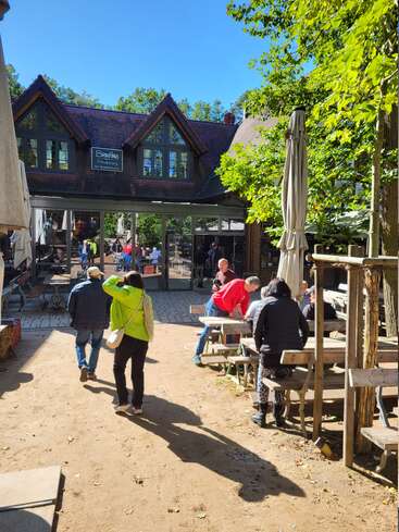 Des personnes profitent d'une journée ensoleillée dans un café en plein air avec des bancs en bois rustiques, des arbres feuillus et un charmant bâtiment en arrière-plan. L'atmosphère est animée et détendue.