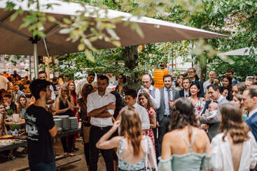 Un grand groupe de personnes se réunit en plein air sous des parasols, écoutant attentivement un orateur. La foule est habillée de façon semi-formelle, ce qui suggère une célébration ou un événement spécial.