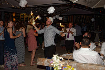 Une scène de fête animée de nuit montre un homme portant un dessert enflammé. Les gens sourient, applaudissent et font la fête sous des guirlandes lumineuses et des décorations suspendues.