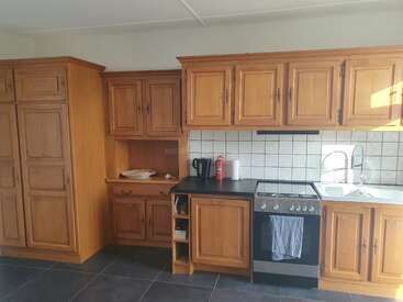 The image shows a kitchen with wooden cabinets, a tiled backsplash, a stove with an oven, a sink, countertop appliances, shelves, and a tiled floor.