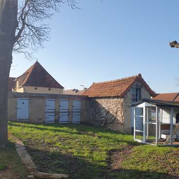 A sunny yard features old stone buildings with tiled roofs, shuttered doors, and windows. A tree and a small glass structure stand on green grassy ground.