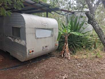 Una vieja caravana desgastada por el paso del tiempo se encuentra bajo un techo metálico junto a un gran depósito de agua, rodeada de exuberantes plantas verdes y bosque, lo que crea un ambiente rústico.