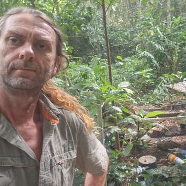 Un hombre con el pelo largo recogido está de pie en un frondoso bosque o jardín. Lleva una camisa de color caqui y mira directamente a la cámara con expresión seria.
