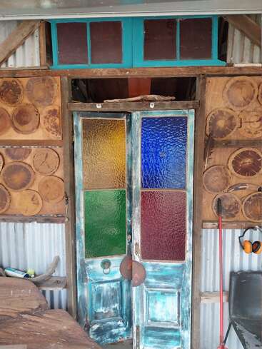 Esta imagen muestra unas puertas dobles rústicas de madera con paneles de vidrieras de colores, rodeadas de rodajas de madera, paredes de chapa ondulada, herramientas y un espacio de trabajo creativo y desgastado por el paso del tiempo.