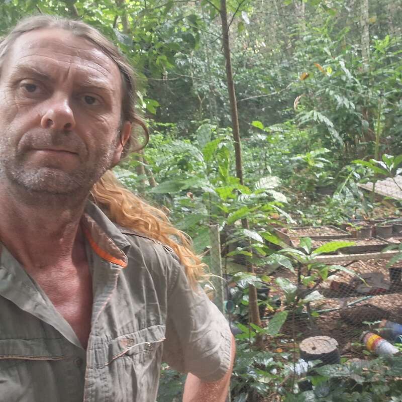 Un hombre con el pelo largo recogido está de pie en un frondoso bosque o jardín. Lleva una camisa de color caqui y mira directamente a la cámara con expresión seria.