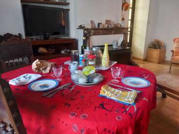 Une table à manger recouverte d'une nappe rouge est dressée pour deux personnes, avec des assiettes, des verres, des produits d'épicerie, des condiments, des couverts et des serviettes de table dans un salon confortable.