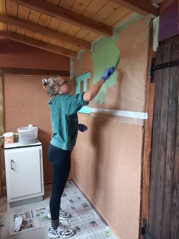 Eine Frau mit grünem Sweatshirt und Brille streicht eine Wand in einem hölzernen Raum hellgrün. Sie steht auf Zeitungen, um den Boden zu schützen, konzentriert und präzise.