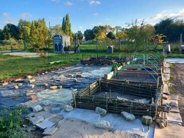 Ein Gemeinschaftsgarten mit erhöhten Holzbeeten, blauem Rahmen, schwarzer Plane und verstreuten Steinen. Ein kleiner blauer Schuppen steht inmitten von grünem Gras, Bäumen und einem sonnigen Himmel.