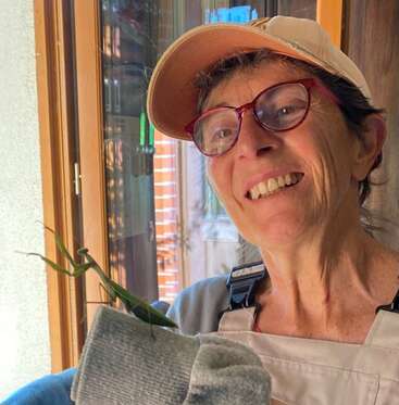 Eine lächelnde Frau mit Mütze und roter Brille hält eine große grüne Gottesanbeterin auf ihrer behandschuhten Hand, während sie in einem Haus an einem sonnigen Fenster steht.