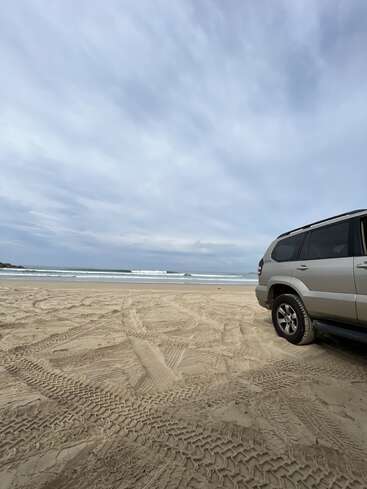 A silver SUV is parked on a sandy beach under a cloudy sky, with tire tracks crisscrossing the sand and gentle waves visible in the distance.